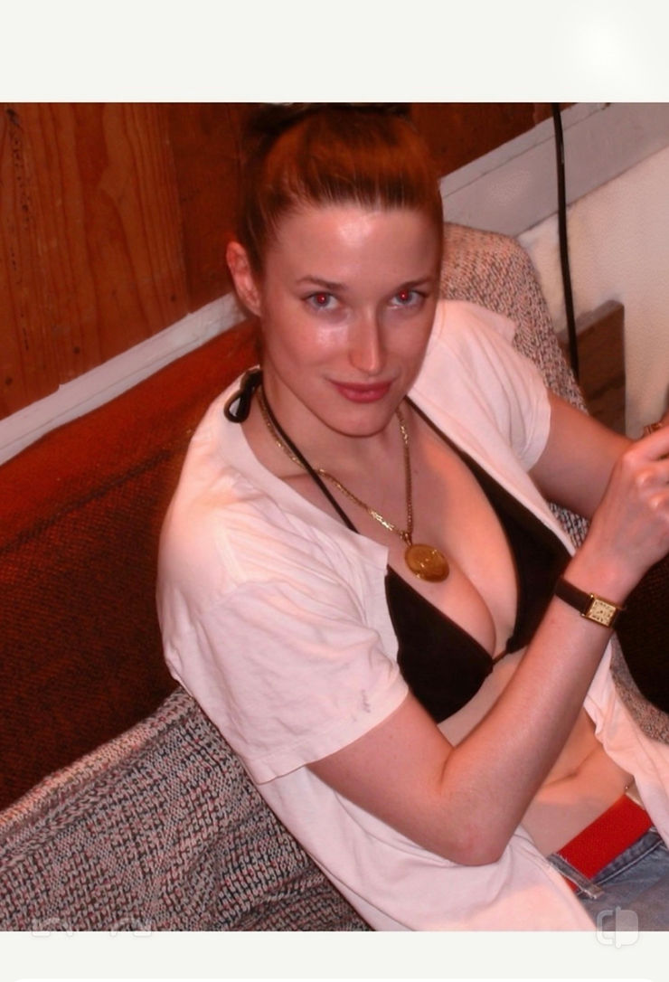 Woman in black bikini top and necklace, sitting on a couch with a wood-paneled background.