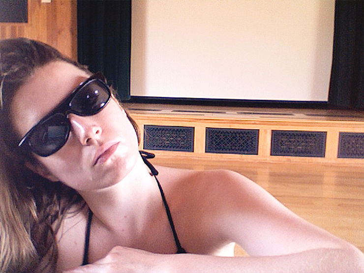 Woman in sunglasses poses indoors, relaxed against a wooden floor.