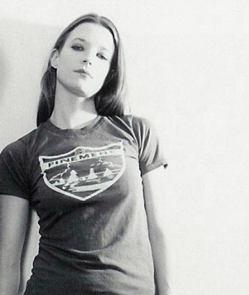 A woman with long hair stands against a light background, wearing a Pine Mtn T-shirt.