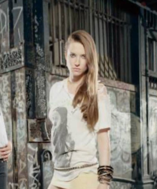 A woman with long hair in a white shirt stands confidently in front of a graffiti-covered wall.